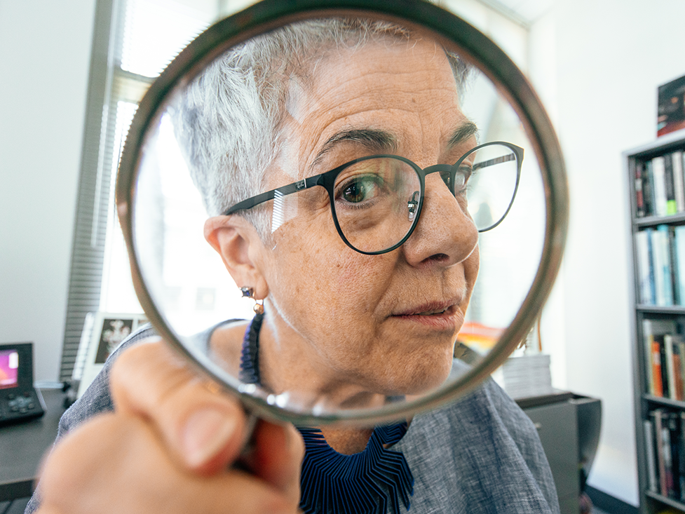 Andrea Pappas in an office with bookshelves peering through a magnifying glass at the camera