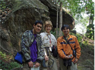 Three people in hiking gear standing in front of a rock formation in a forest.