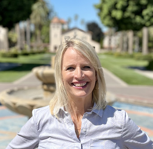 A person smiling outdoors with trees and a path in the background.