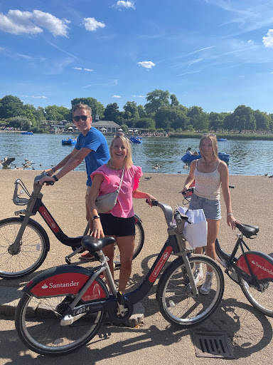 Three people with bicycles standing on a lakeshore with trees in the background.