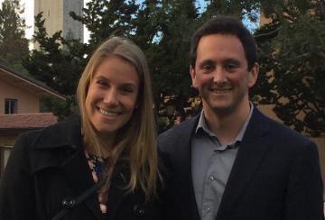 Two people smiling outdoors with buildings and trees in the background.