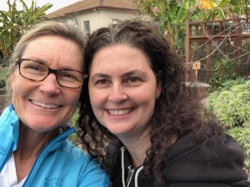 Two people smiling outdoors with trees and buildings in the background.