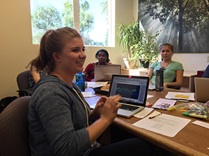 Alt text: Students engaged in a discussion around a table, one displaying research on a laptop.