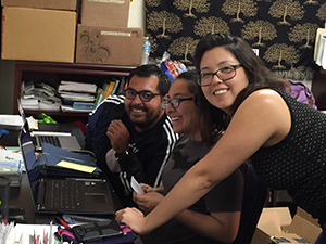 Alt text: Three students smiling and posing indoors with papers and books in the background.