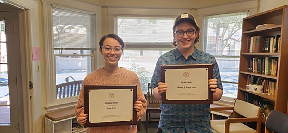 2 students holding award plaques in a bay windowed house