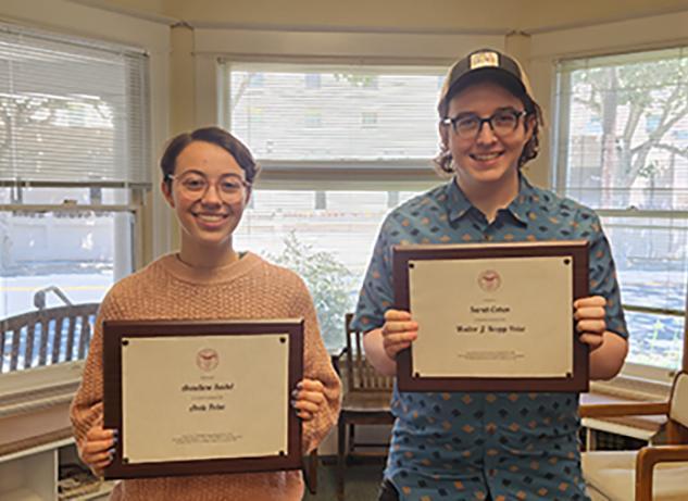 2 students holding award plaques in a bay windowed house