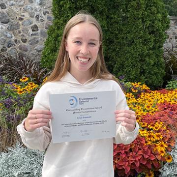 A person holding an award certificate in front of colorful flowers and greenery.