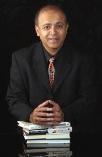 A person with several stacked books, wearing a suit and tie, with black background.