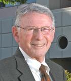 Older man wearing glasses and a suit, smiling outside a building.