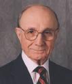 A person wearing glasses, a suit, and a tie with a neutral expression.