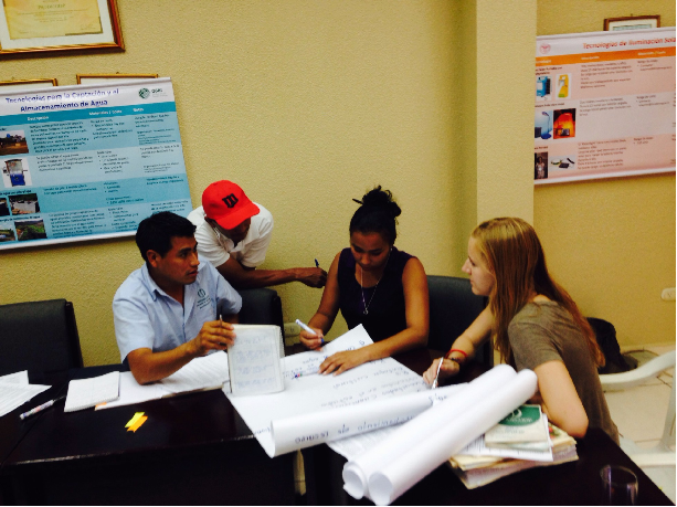 Four students collaborating at a table with documents and charts on the walls.