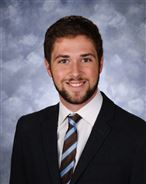 A man in a suit and tie smiling against a cloudy background.