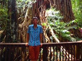 A person posing in front of a large tree within a lush forest.