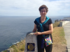 Person standing beside a marker near the coast with the sea and sidewalk.