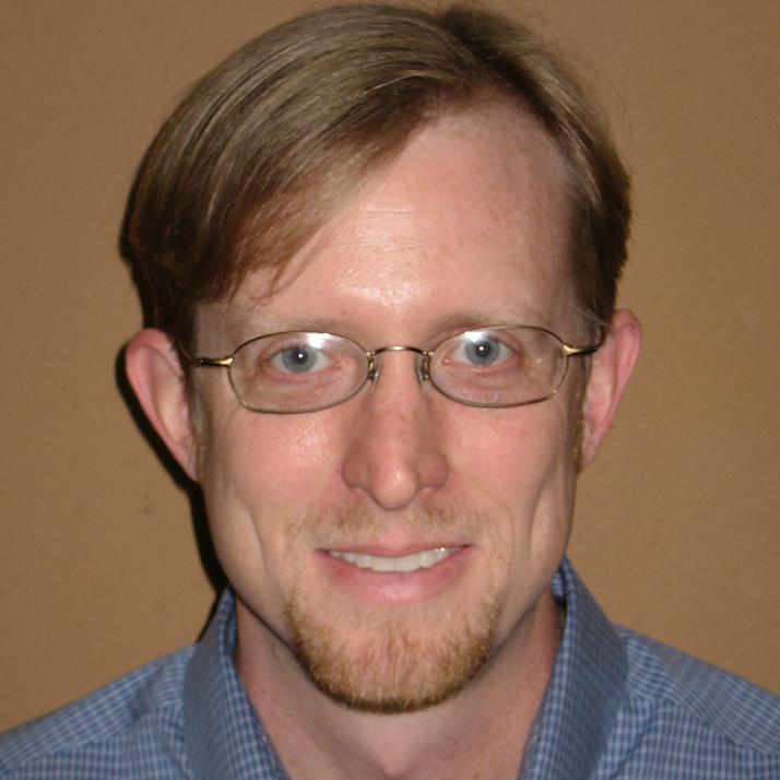 Alt text: Person wearing glasses and a blue shirt against a brown background.