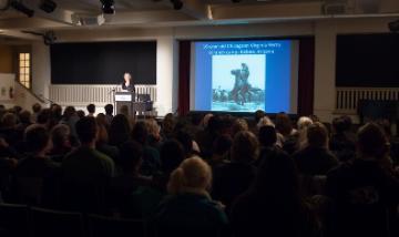 A speaker presenting to a large audience with a slideshow on display.