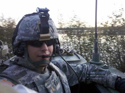 A soldier in camouflage, wearing a helmet and sunglasses, with foliage in the background.