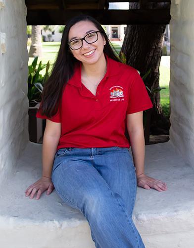 Person in a red polo shirt and blue jeans sitting outside and smiling.