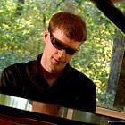 A person wearing sunglasses playing a grand piano outdoors.