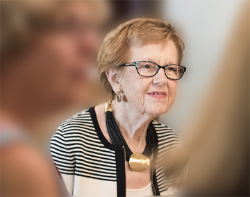 An elderly woman with glasses smiles, slightly blurred people in the foreground.