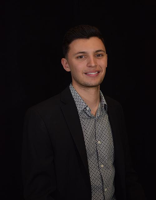 A person in a dark suit with a patterned shirt smiles against a black background.