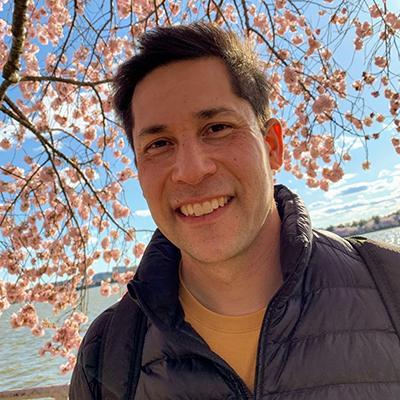 A person smiling in front of blossoming cherry trees.