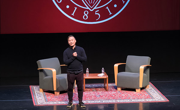 A person speaking on stage with two chairs and a table.