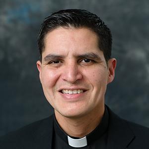 A smiling person wearing a clerical collar and a black jacket.