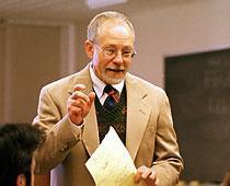 A person speaking in a classroom holding papers.