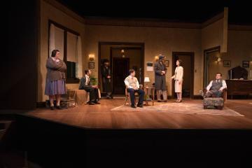 Theater stage scene with six actors amidst vintage furniture and decor.