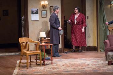 A stage scene with two people standing and conversing in a living room setting.