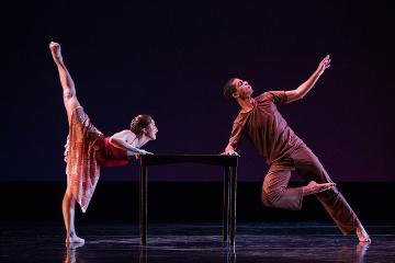 Two dancers perform around a table on stage.