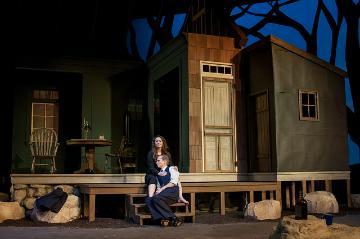 Person sitting on stage in front of a rustic wooden cabin set.