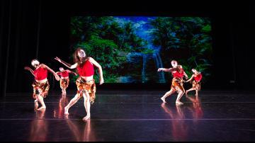 Dancers in red costumes performing on stage with tropical backdrop.