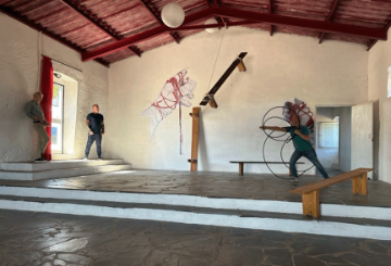 2 men stand on the left and 1 man stands on the left of a large room with cream walls, a wooden roof and grey tiled floor.  There's a bench on the right and modern art on the far wall.