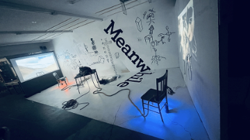 Artist studio with black and white art on the walls, lit by a digital project on a table and screen hung on a wall, with cables on the floor and 2 chairs