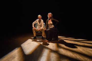 A man and a woman sitting on chairs next to each other having a conversation in a darkened room lit only by sunlight through the window