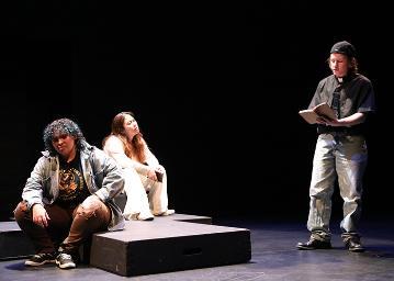 2 women sitting on a low stage looking up at a standing man reading from a book