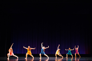 Dancers in colorful outfits performing on stage with a dark background.