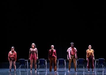 5 students each standing behind a chair desk combo in the middle of the stage