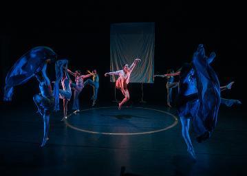 Female dancer in a white shirt and brown shorts in a tuck jump surrounded by 4 dancers on each side with material as waves