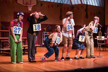 Participants on stage at a spelling bee competition, making expressive gestures.