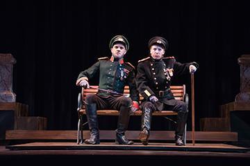 Two actors in formal attire sit on chairs on stage in 