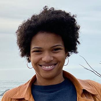 A person smiles, wearing a brown jacket and blue shirt outdoors.