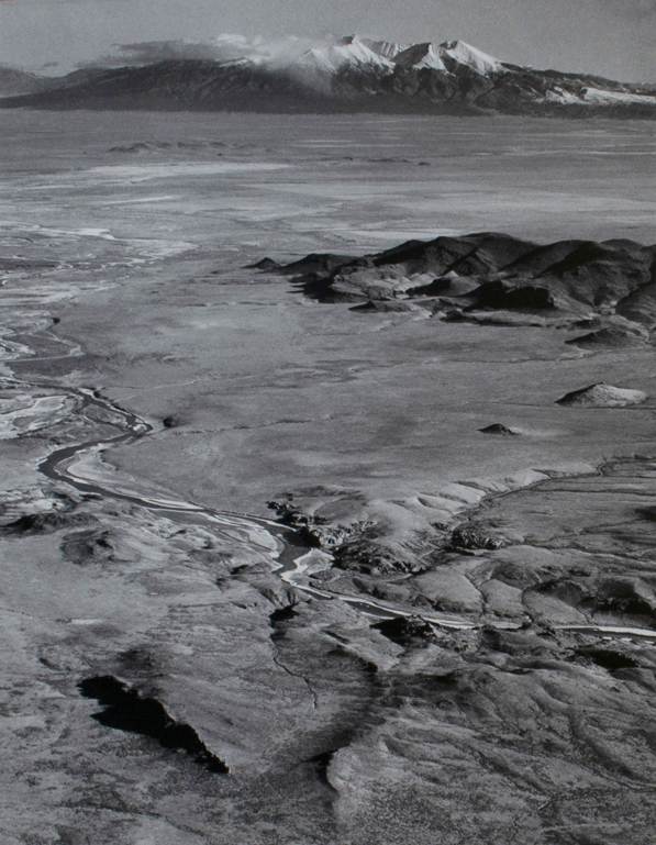 Black and white photograph of aerial view of river valley and mountains. River runs diagonally in the lower part. Snow capped mountains in the distance.