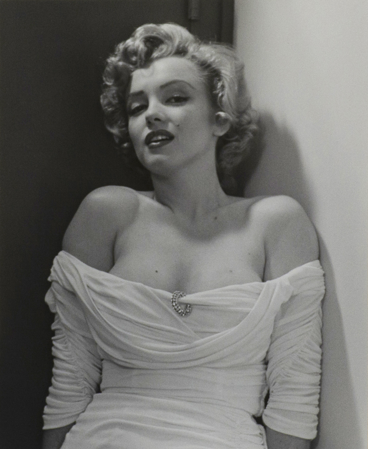 Black and white photograph of Marilyn Monroe looking directly to the viewer. Figure is leaning back and wearing a white dress with a brooch in the center, dress sleeves worn below the shoulder.