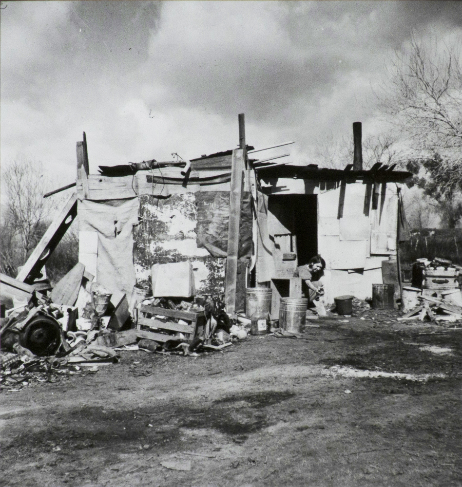 Black and white photograph of shack made of boards, tar paper and other salvaged materials. Debris and trash around building, a figure sits in the doorway. Trees and fields in the background.