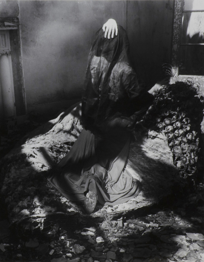 Black and white photograph of figure draped with a sheer veil over their head with their left hand on top. Left side of image is a Greek ionic column. On right side of image is a window frame.