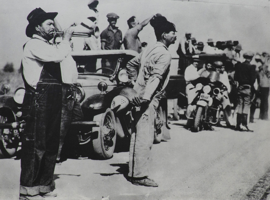 Black and white photograph of of figures standing in front of car which is leading caravan. Man to left in dark hat wearing coveralls is blowing a bugle. Police officer on motor bike in center of picture. More figures standing around in other cars in rear.