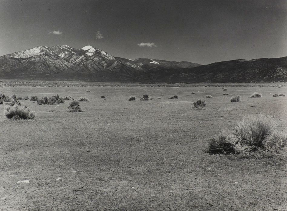 Black and white photograph of an open field land with sagebrush bushes throughout the field. Partially snow covered mountain range in background. Three small puffs of clouds above snow peaks.